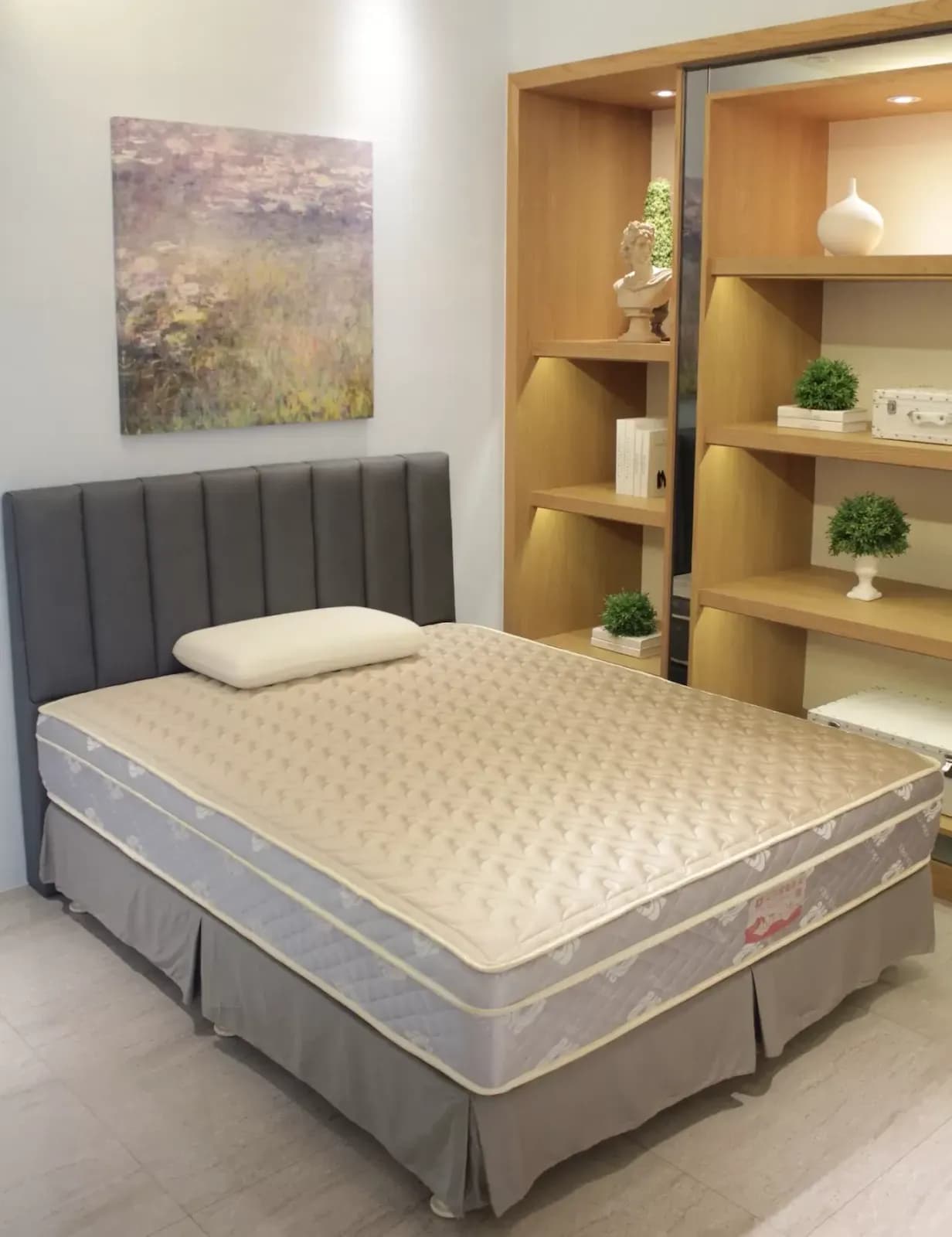 臥室中擺放米色點狀床墊與灰色裙襬床架，深灰色軟墊床頭靠牆，後方是木質展示櫃與裝飾品。