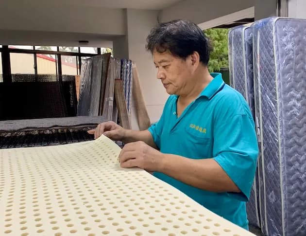 一名穿藍綠色上衣的男子站在床墊旁，雙手檢查米色泡棉表面，背景可見直立收納的多張床墊。