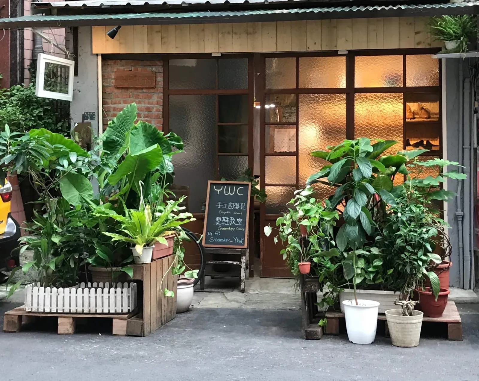 一間小店入口被多盆綠色觀葉植物包圍，中央有玻璃門與黑板，黑板上寫著 WC 及中文文字。