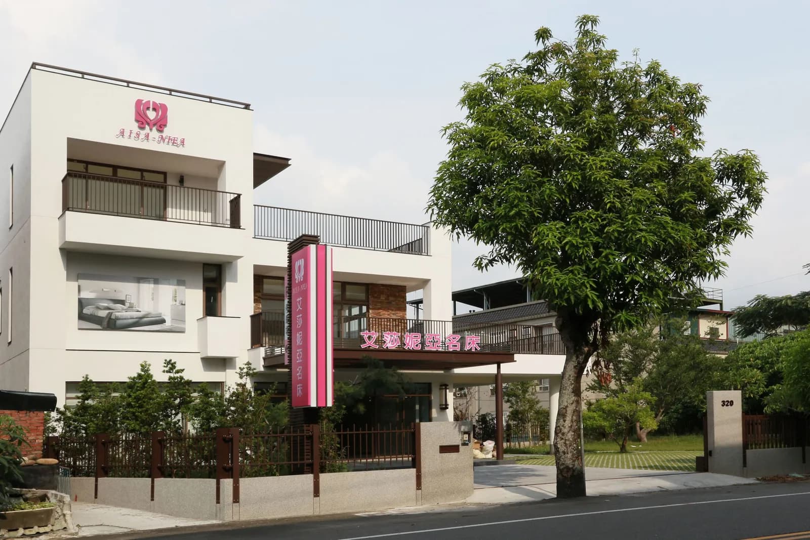 白色現代建築外觀設有陽台與落地窗,入口前有粉紅色中文招牌、圍牆、樹木及臨路綠地。