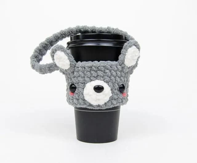 黑色杯子套著灰色毛線鉤織杯套,正面有白色口鼻、黑色鼻子與眼睛,兩側連著圓耳朵。