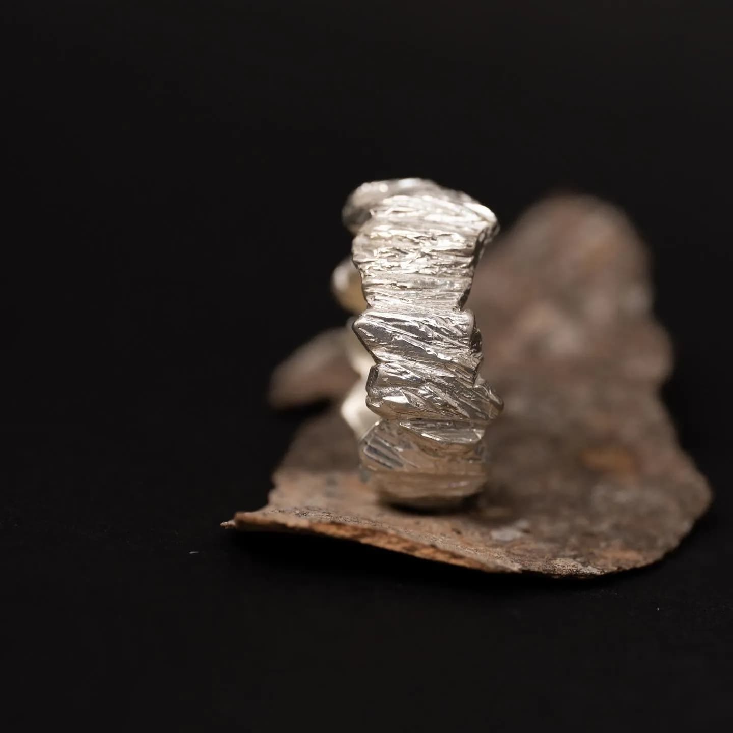 黑色背景中央,一枚銀色立體戒指直立在褐色粗糙薄片上,戒面呈現層疊皺褶與不規則輪廓。