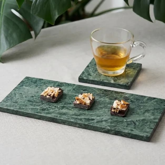 透明玻璃茶杯盛著金黃色液體,放在深綠色大理石杯墊上,前方大理石托盤排列三塊堅果點心,背景有大片綠葉。