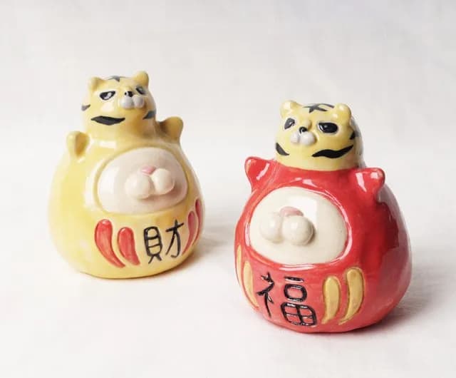 白色背景前擺著黃色與紅色兩個老虎造型圓陶偶,腹部有凸起裝飾,並繪有黑色中文字樣。