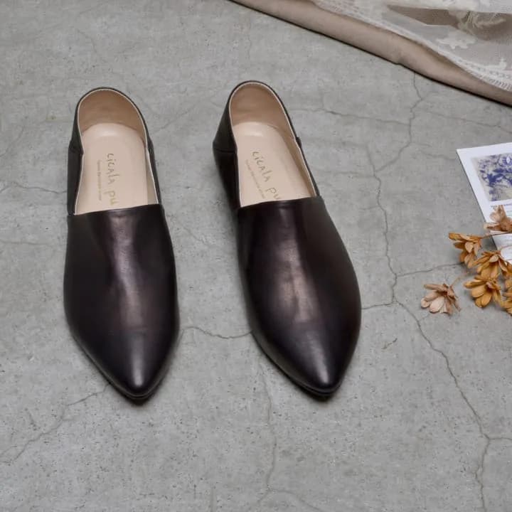 一雙深棕色尖頭皮革穆勒鞋並列放在灰色裂紋地面,鞋內露出淺棕色鞋墊,右側散落乾燥花。