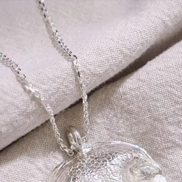 銀色細鍊垂掛在米白色布料上,中央連接一枚圓形吊墜,表面呈細密凹凸紋理。
