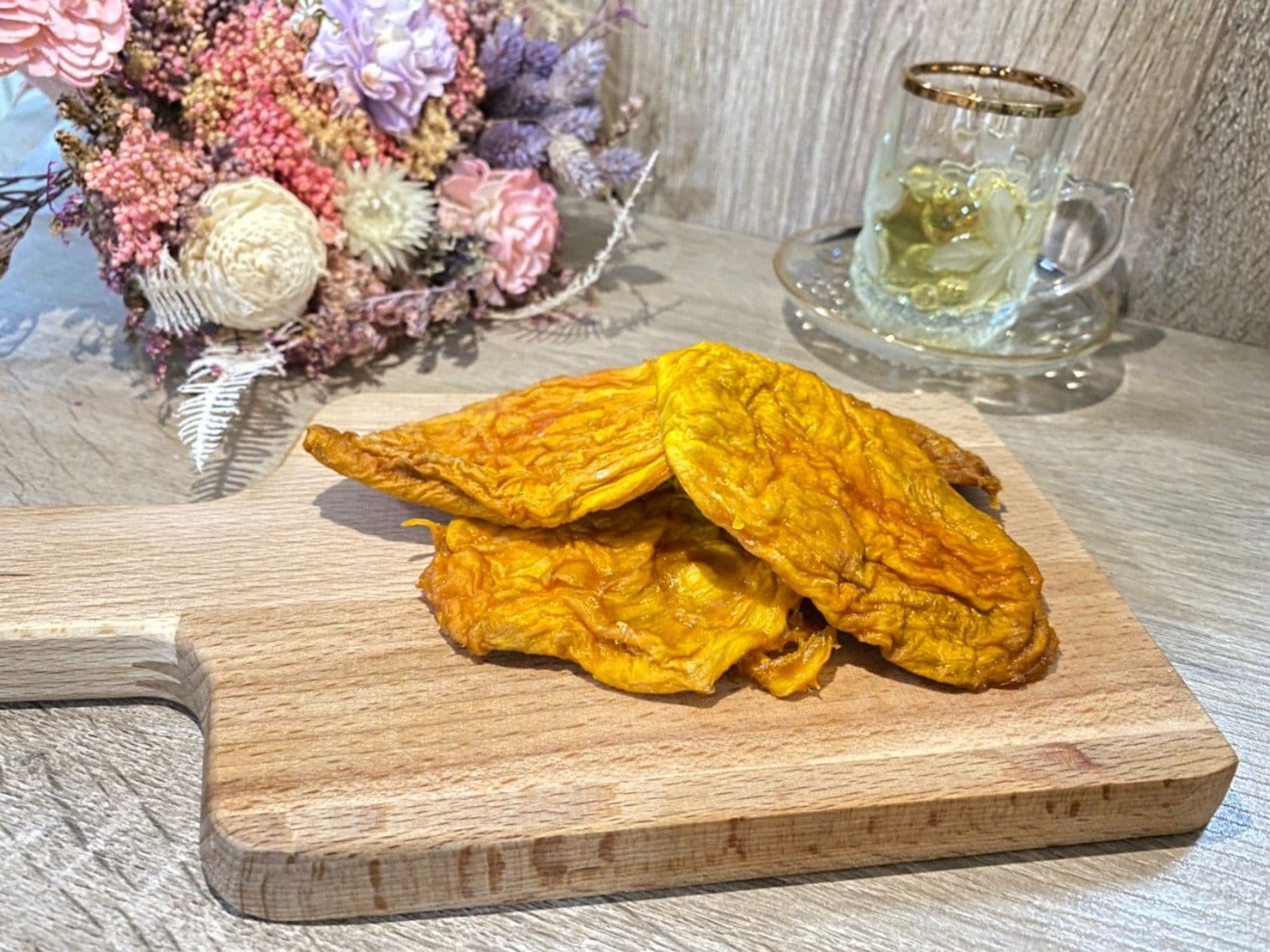 Dried mango from 日青芒果 displayed on a wooden cutting board with a tea cup and floral decoration in the background.