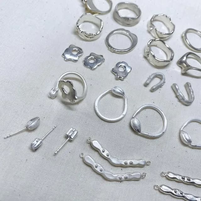 多種銀色金屬飾品零件散放在淺灰色布面上,包括圓環、花形片與細長鏈節。