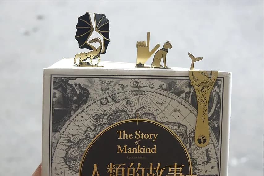 一本印有古地圖與「The Story of Mankind」字樣的書立在畫面中央,書頂擺放黑金葉片、人物與動物造型金屬裝飾。