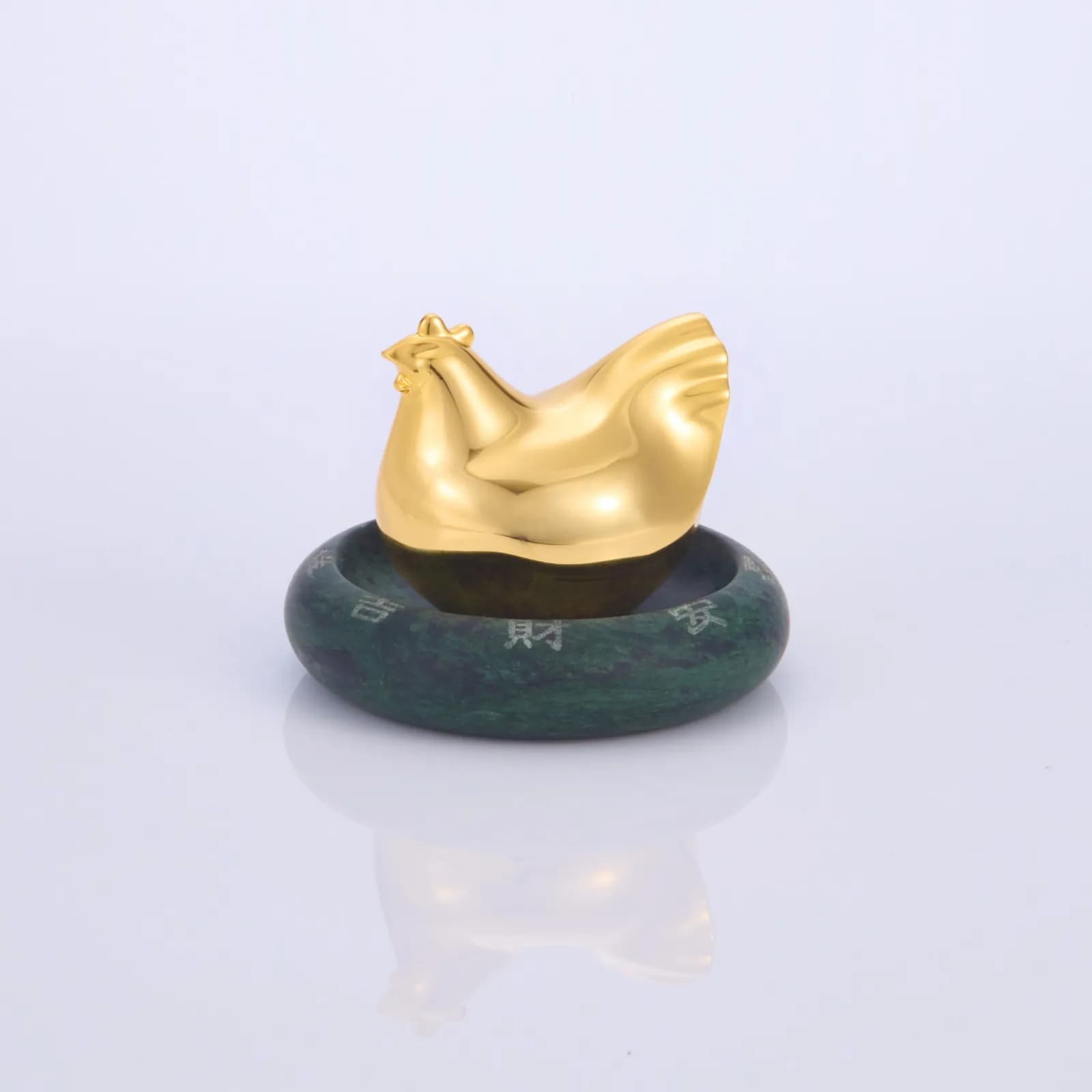 白色背景中央擺放金色雞形雕像,下方是深綠色圓形底座,底座表面刻有淺色文字,地面映出淡淡倒影。