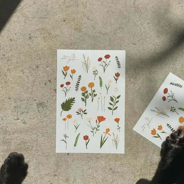A sheet of botanical illustrations with colorful flowers and leaves on a textured gray surface, with part of another illustrated sheet visible at the side.